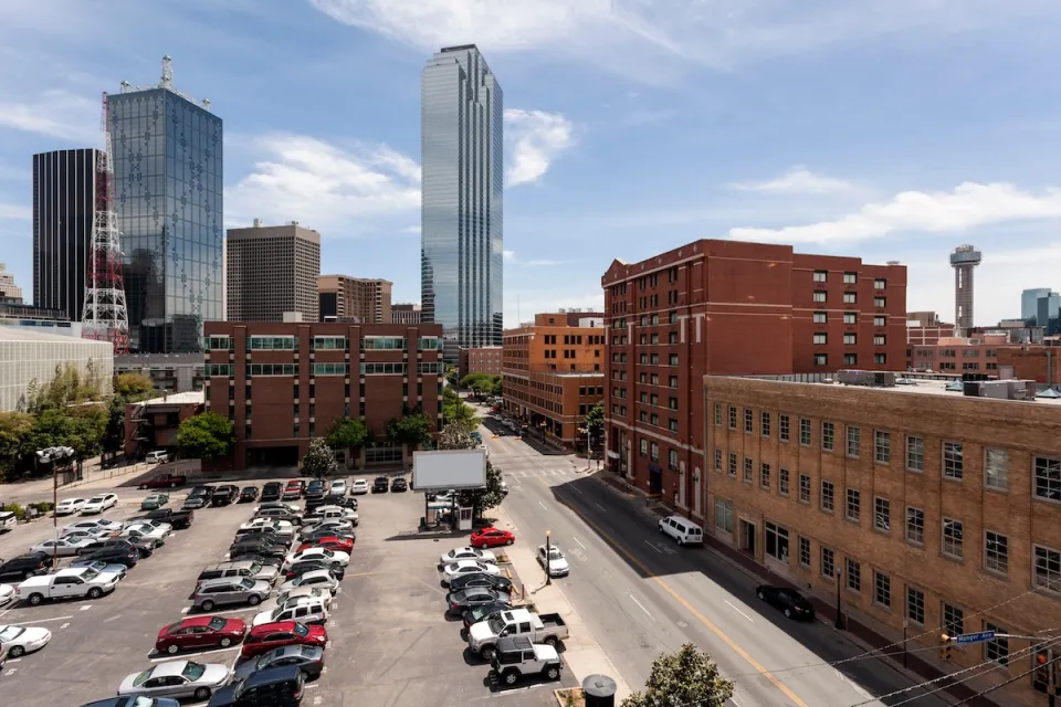 View of mostly full parking lot next to multi-story red brick buildings with modern glass skyscraper in background in downtown Dallas, Texas.